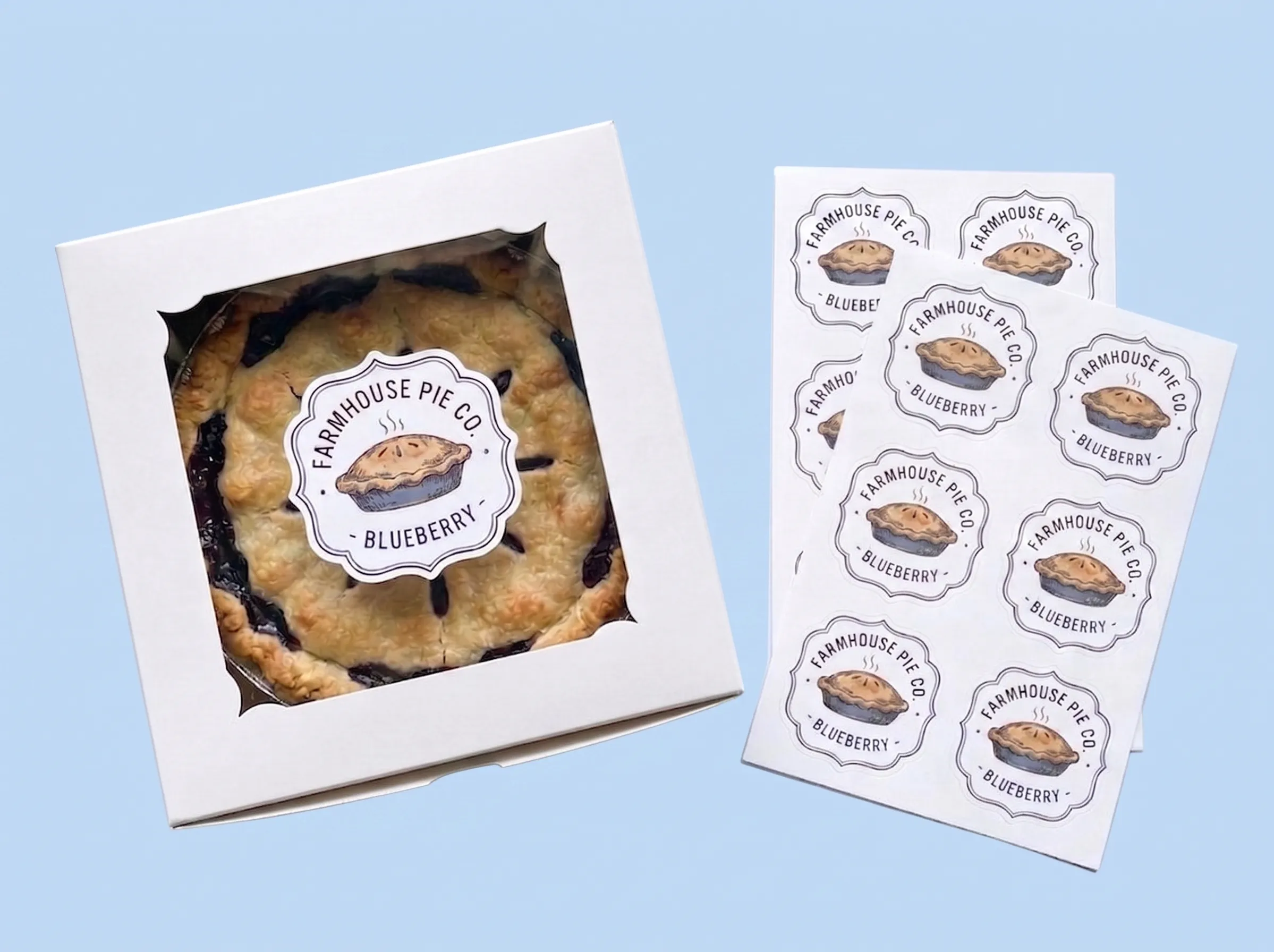 A top-down view showcasing custom die-cut pie labels ready for bakery application. On the left, a freshly baked blueberry pie is packaged in a white window box, featuring a custom shape label applied directly to the clear window. On the right, two printed sheets of identical custom labels are displayed, illustrating how the stickers are delivered on sheets for quick and easy peeling during production.
